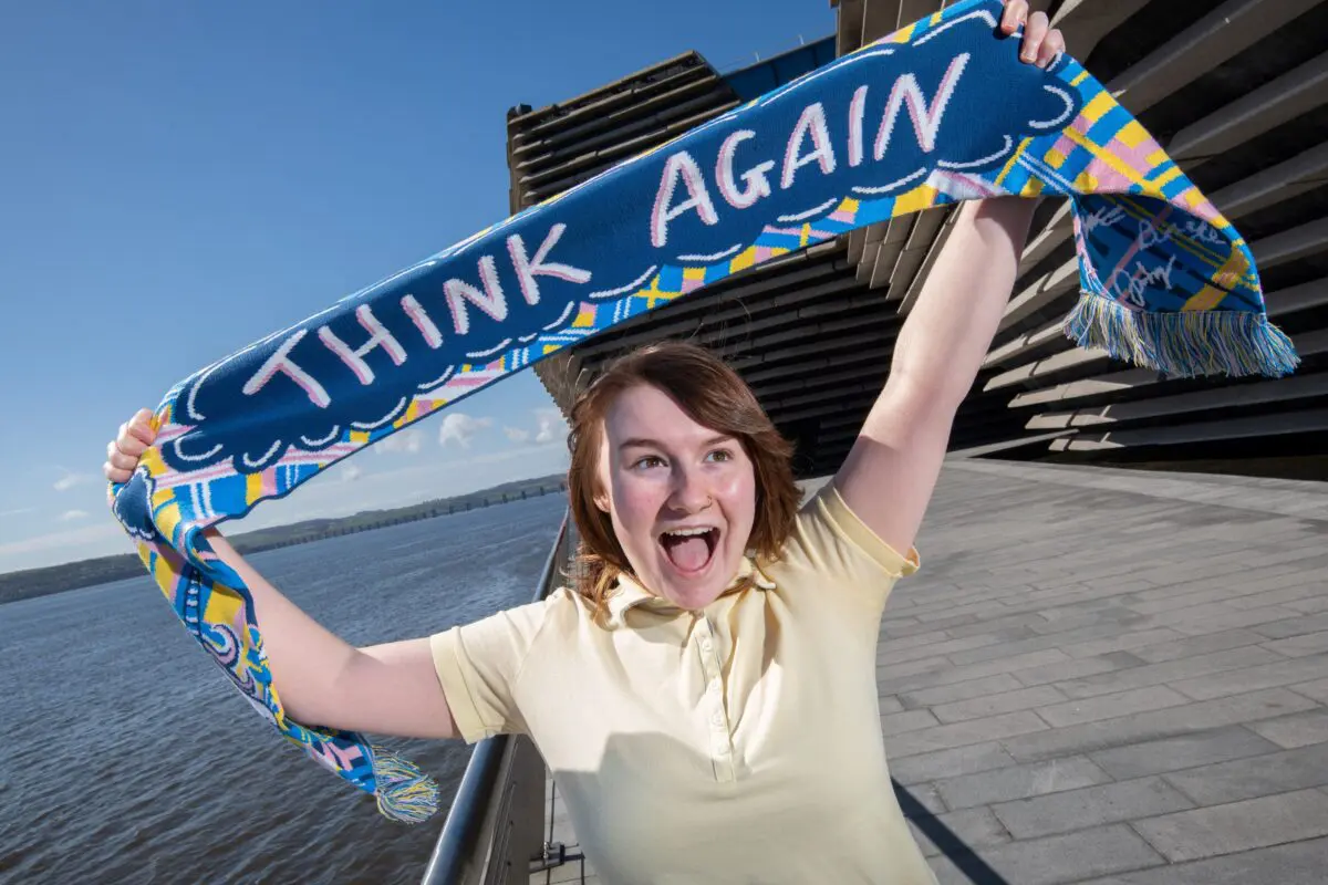 Person cheering, holding up a blue scarf reading "THINK AGAIN" beside a waterfront and stepped concrete building.