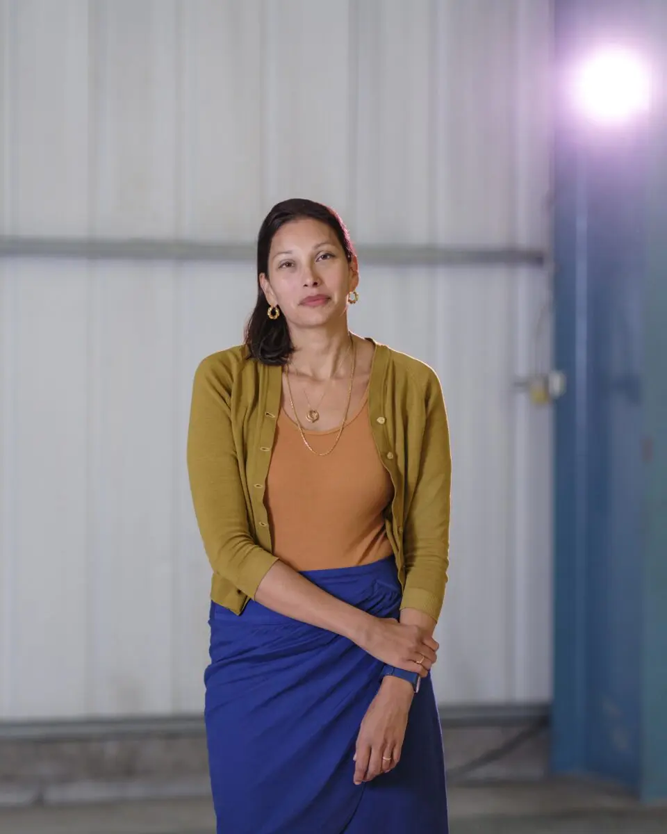 Woman against a pale wall wearing a mustard cardigan, orange top, blue skirt and gold jewelry, hands folded at waist.