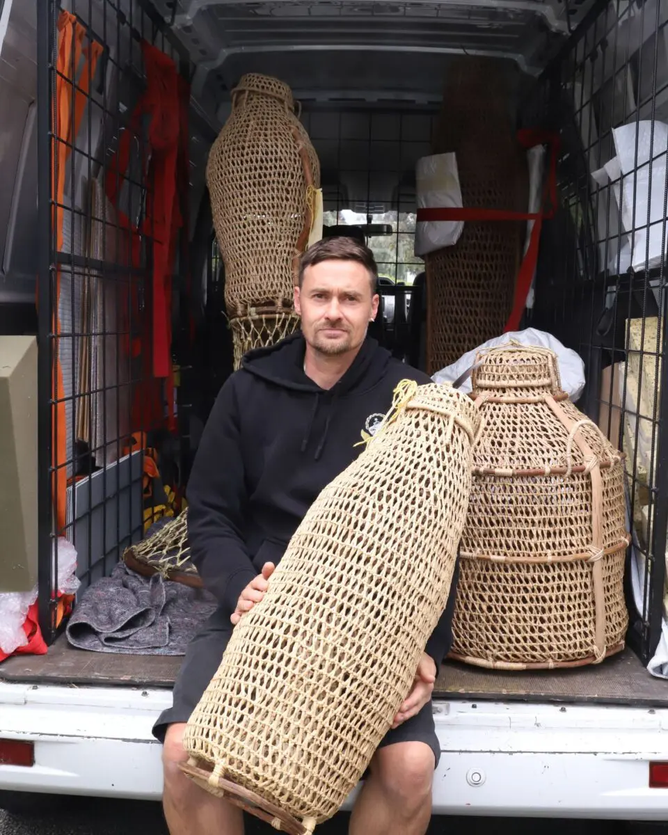 Man sitting in the back of a van holding a large woven fish trap, with wicker baskets stacked behind him.