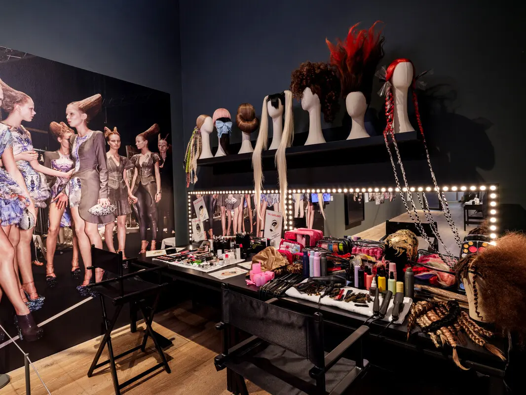 Fashion backstage dressing table with lit mirror, wigs on mannequin heads, makeup and hairstyling tools.