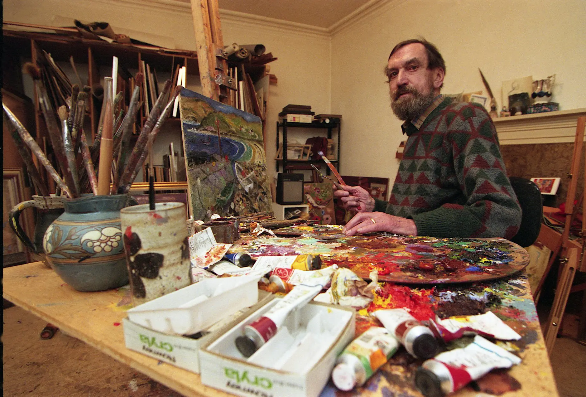 Bearded man seated at a messy studio table, holding brushes beside a small landscape and a paint-splattered palette.