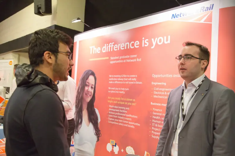 Two men speaking at a trade-show booth with a large red promotional backdrop featuring a woman's photo.