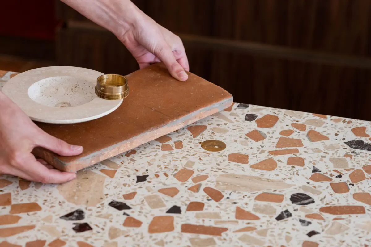 Hands placing a terracotta tile and a small stone sink bowl with brass ring on a terrazzo countertop.