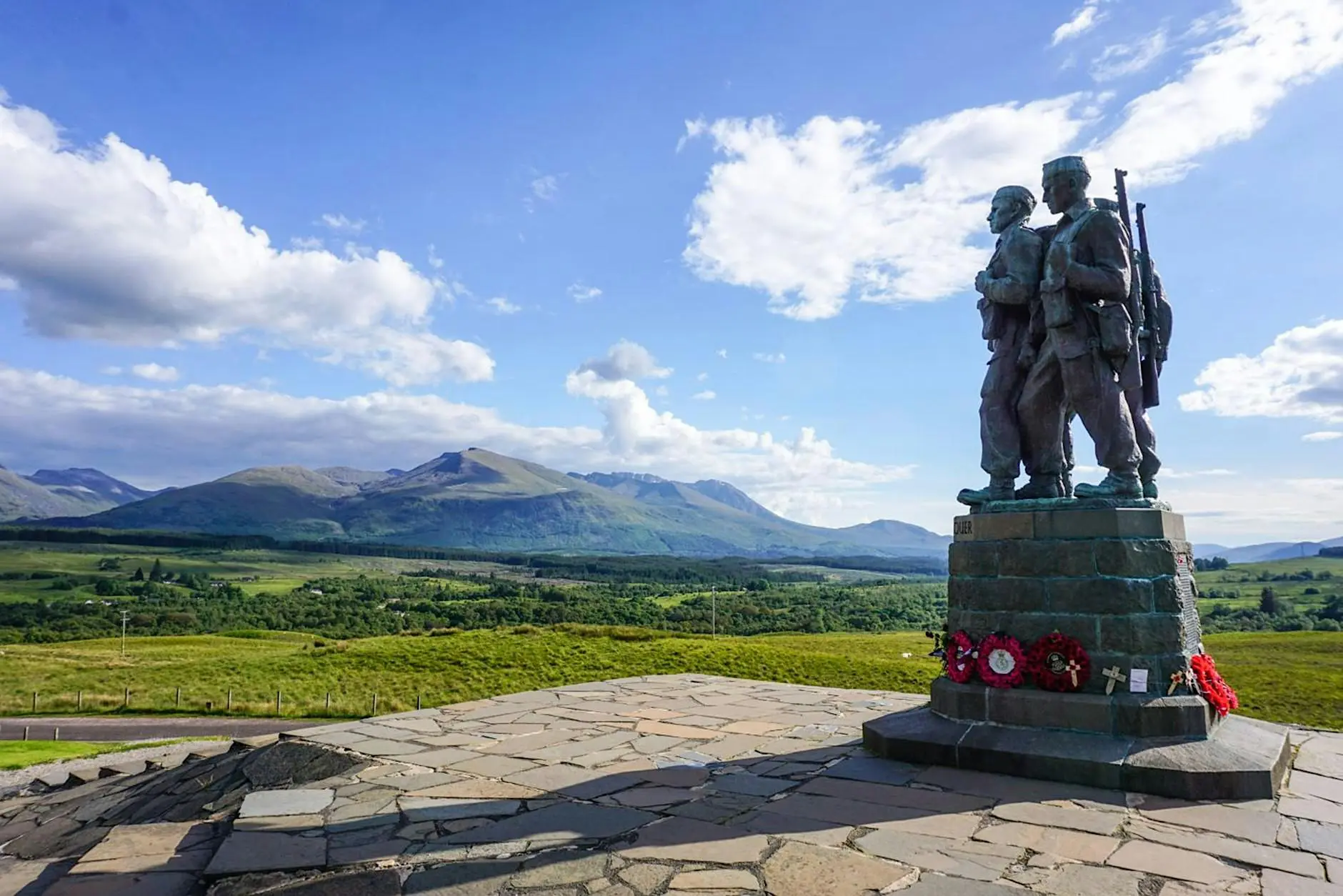 Bronze soldier monument with wreaths on a stone plinth overlooking a green valley and distant mountains under a blue sky.