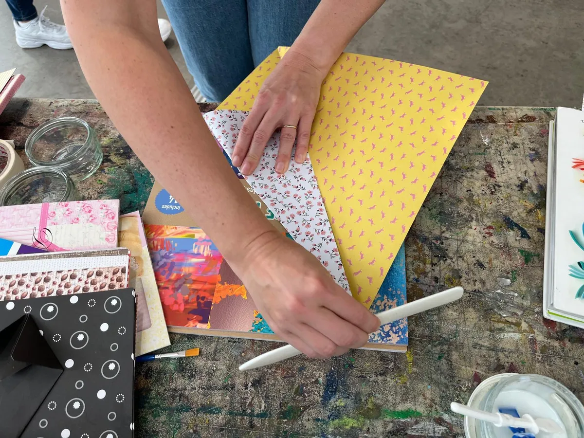 Hands folding yellow and floral patterned paper with a bone folder on a paint-splattered craft table