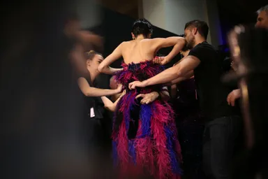 Model seen from behind in a purple and pink feathered gown as several stylists adjust it backstage.