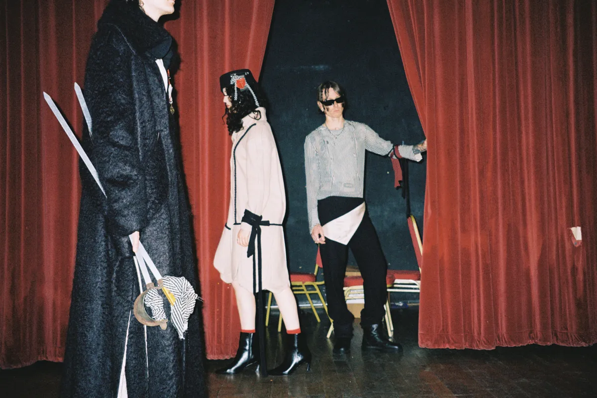 Three people in eclectic costumes stand by red stage curtains, one pulls them open while another holds props.
