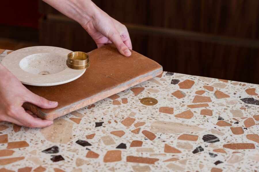 Hands placing a terracotta tile and a small stone sink bowl with brass ring on a terrazzo countertop.