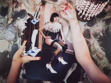 Woman in black corset and stockings posed on a stool, surrounded by hands placing white high heels and decorative lace