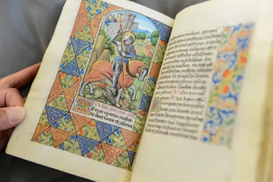 Hands holding illuminated manuscript with colorful mounted knight illustration, ornate floral borders and Gothic text.