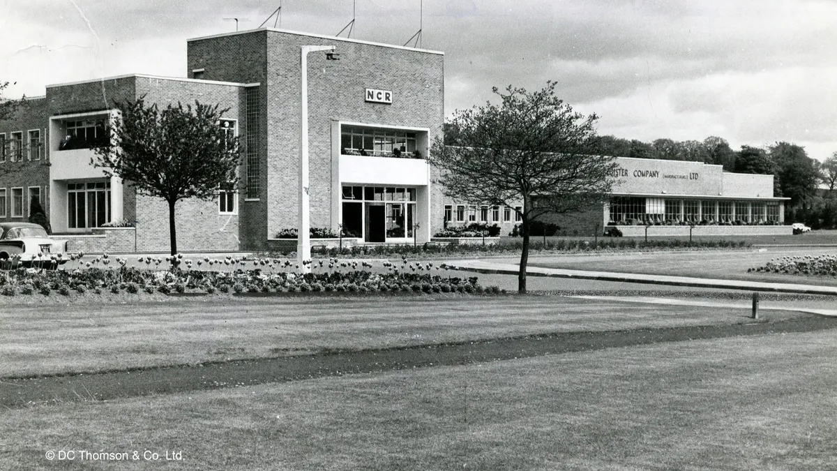 Black-and-white midcentury NCR office and factory building with manicured lawn, trees, and a parked car.