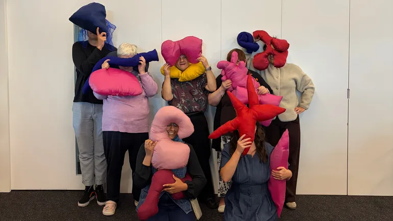 Group of eight people posing against a white wall, each holding colorful abstract-shaped cushions that cover their faces.